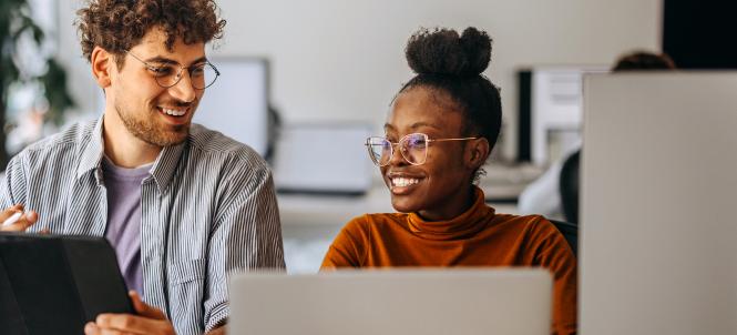 A man and a woman are sitting at a desk with open laptops in from of them. They are looking at one another and smiling.