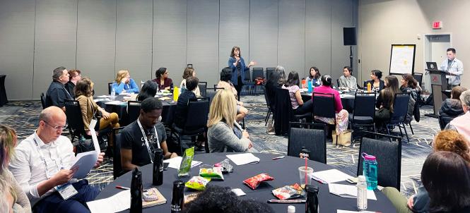 A woman talks through a microphone to a room full of people at tables during a Code for America session.