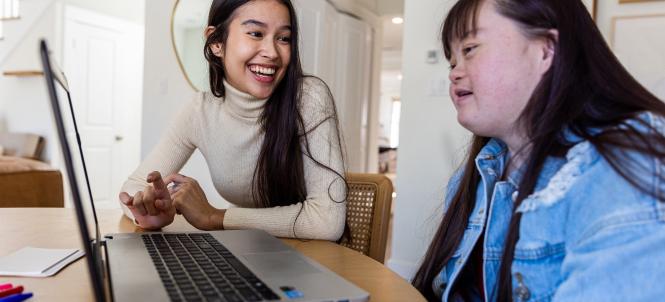 Two women sit at a table looking at a laptop.