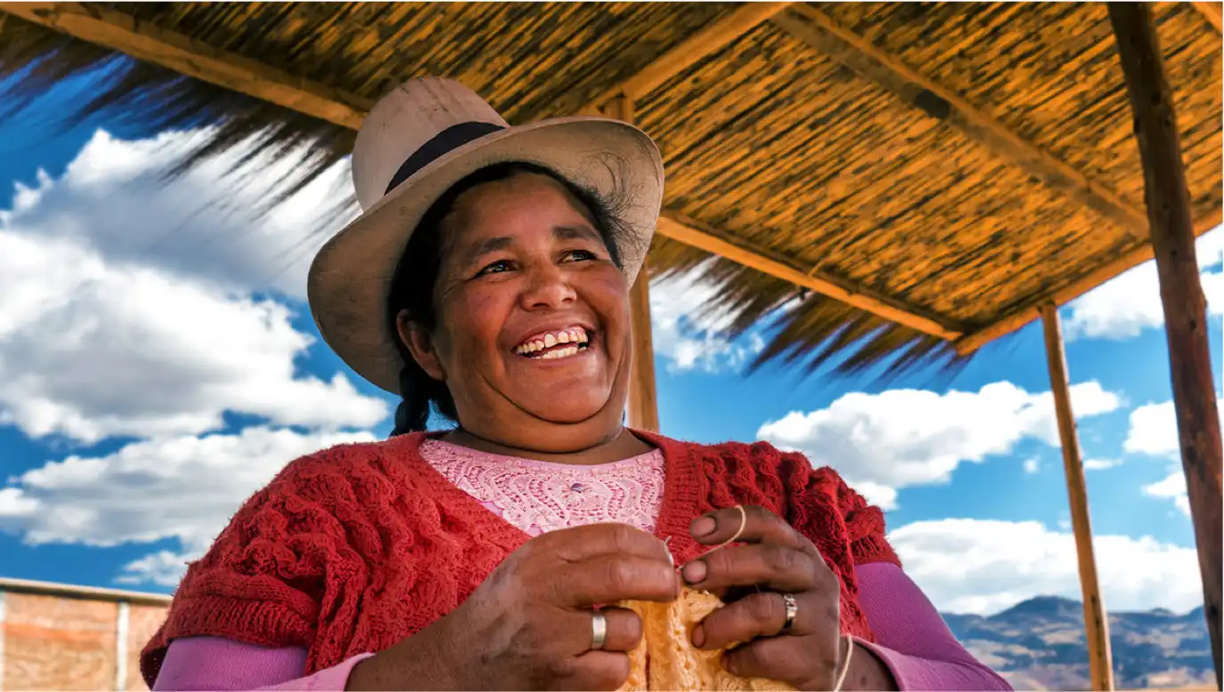 An indigenous woman smiling while knitting outside underneath a thatch-covered roof.